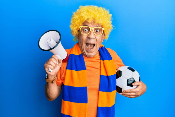 Hombre hispano mayor hooligan fútbol animando juego celebración de pelota y megáfono celebrando loco y sorprendido por el éxito con los ojos abiertos gritando emocionado.  - Foto, Imagen