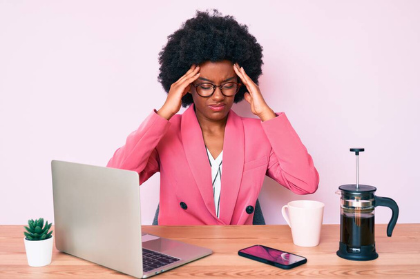 Молода афроамериканська жінка, яка працює за столом за комп'ютерним ноутбуком з руками на голові, головний біль, тому що стрес. мігрень..  - Фото, зображення