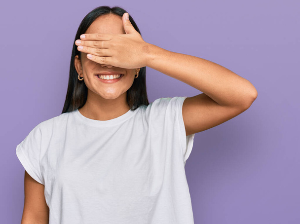 Jovem mulher asiática vestindo casual t-shirt branca sorrindo e rindo com a mão no rosto cobrindo os olhos para surpresa. conceito cego.  - Foto, Imagem