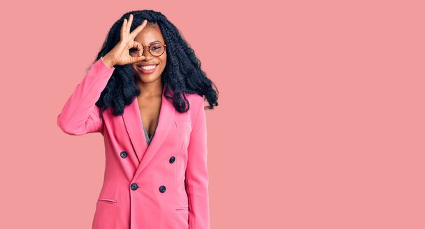 Mulher americana africana bonita vestindo jaqueta de negócios e óculos sorrindo feliz fazendo ok sinal com a mão no olho olhando através dos dedos  - Foto, Imagem