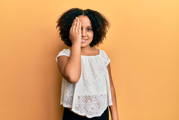 Young little girl with afro hair wearing casual clothes covering one eye with hand, confident smile on face and surprise emotion.  - Фото, зображення