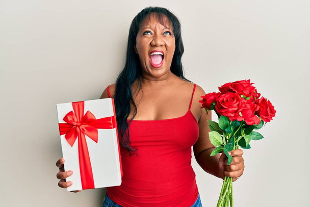 Mulher afro-americana de meia-idade segurando presente de aniversário e buquê de flores irritadas e loucas gritando frustradas e furiosas, gritando com raiva olhando para cima.  - Foto, Imagem