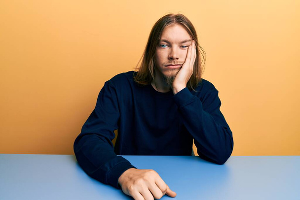 Handsome caucasian man with long hair wearing casual clothes sitting on the table thinking looking tired and bored with depression problems with crossed arms.  - Foto, Bild