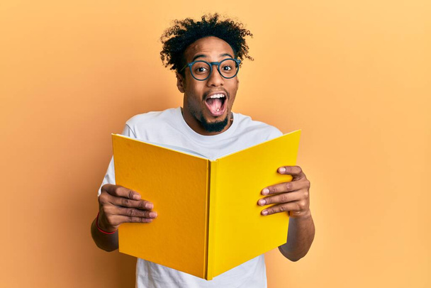 Молодой африканский американец с бородой читает книгу в очках, празднуя безумие и поражаясь успеху с открытыми глазами, крича взволнованно.  - Фото, изображение
