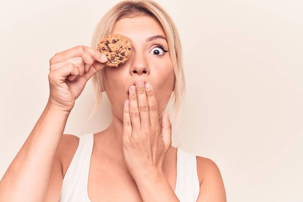 Mladá krásná blondýnka drží sladkou čokoládovou sušenku přes oko zakrývající ústa rukou, šokovaná a vystrašená z omylu. Překvapený výraz - Fotografie, Obrázek