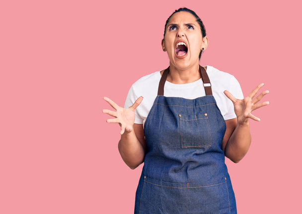 Young beautiful brunette woman wearing apron crazy and mad shouting and yelling with aggressive expression and arms raised. frustration concept.  - Foto, Imagen
