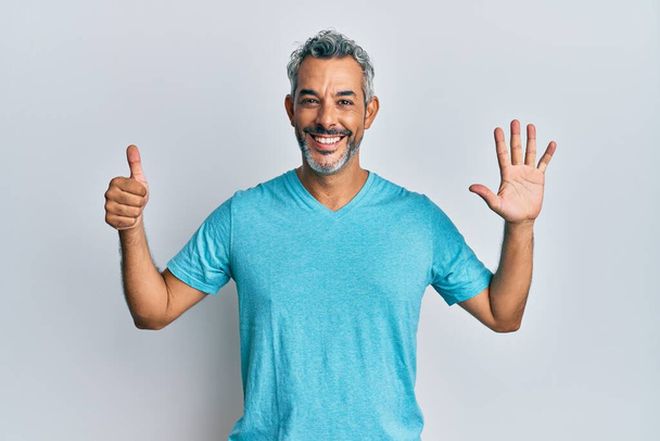 Středního věku šedovlasý muž na sobě ležérní oblečení ukazuje a ukazuje nahoru s prsty číslo šest a zároveň se usmívá sebevědomě a šťastně.  - Fotografie, Obrázek