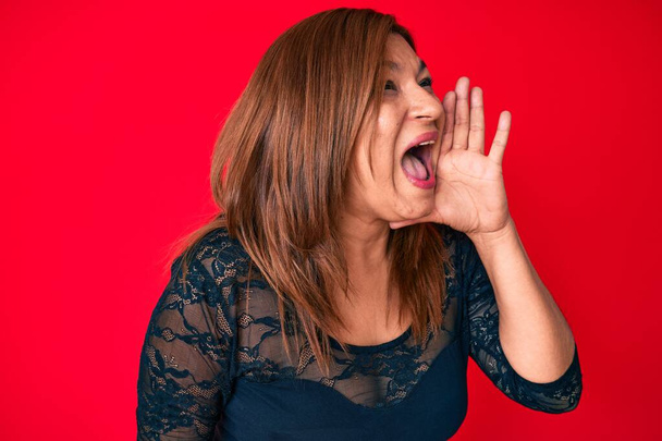 Middle age brunette hispanic woman wearing elegant clothes over red background shouting and screaming loud to side with hand on mouth. communication concept.  - Photo, Image