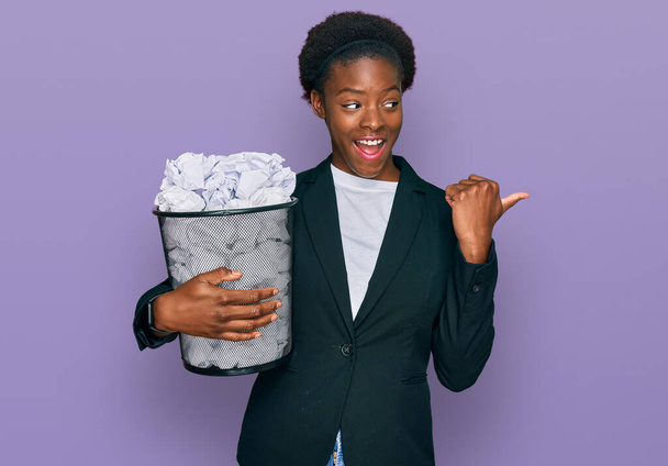 Junge Afroamerikanerin hält Papierkorb voller zerknüllter Papiere und zeigt mit dem Daumen zur Seite, lächelt glücklich mit offenem Mund  - Foto, Bild