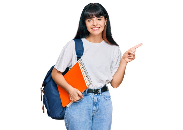 jong hispanic meisje dragen student rugzak en vasthouden boek glimlachen gelukkig wijzend met hand en vinger naar de zijkant  - Foto, afbeelding