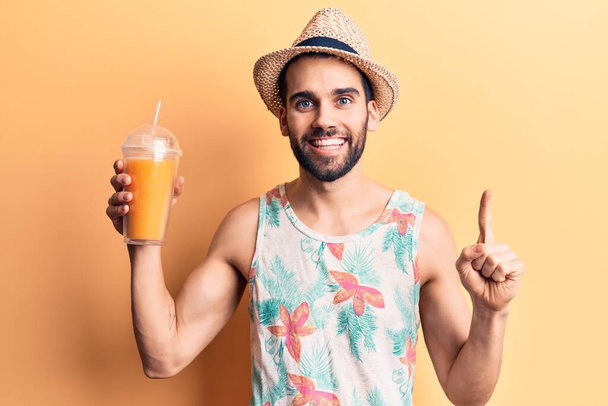 Νεαρός όμορφος άντρας με γενειάδα φορώντας καλοκαιρινό καπέλο και μπλουζάκι πίνοντας χυμό πορτοκάλι χαμογελώντας με μια ιδέα ή ερώτηση δείχνοντας δάχτυλο με χαρούμενο πρόσωπο, νούμερο ένα  - Φωτογραφία, εικόνα