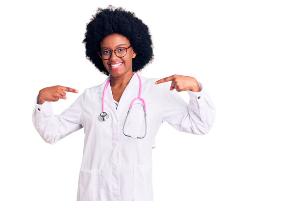Fiatal afro-amerikai nő orvosi kabátot és sztetoszkópot visel, magabiztosan néz ki, mosollyal az arcán, ujjaival büszkén és boldogan mutogat magára..  - Fotó, kép