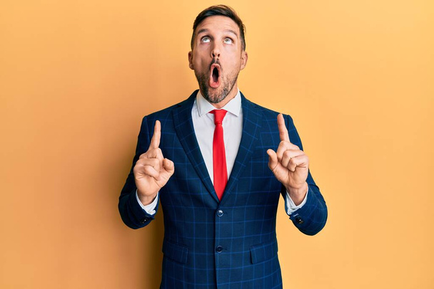 Handsome man with beard wearing business suit and tie amazed and surprised looking up and pointing with fingers and raised arms.  - Foto, Imagem