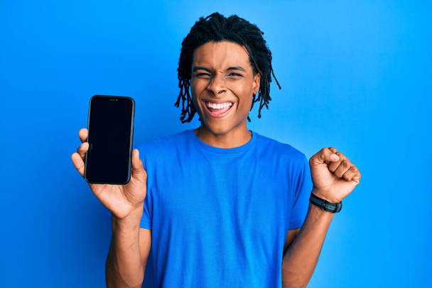 jeune homme afro-américain tenant smartphone montrant écran blanc criant fier, célébrant la victoire et le succès très excité avec le bras levé  - Photo, image
