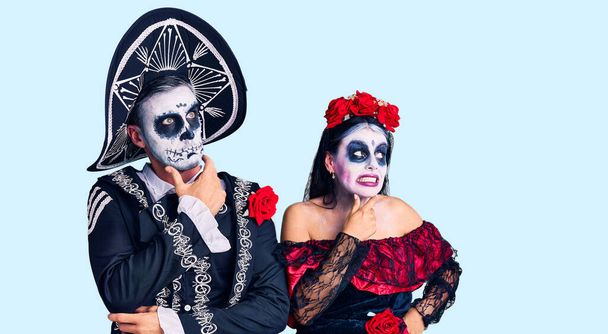 Young couple wearing mexican day of the dead costume over background thinking worried about a question, concerned and nervous with hand on chin  - Fotografie, Obrázek