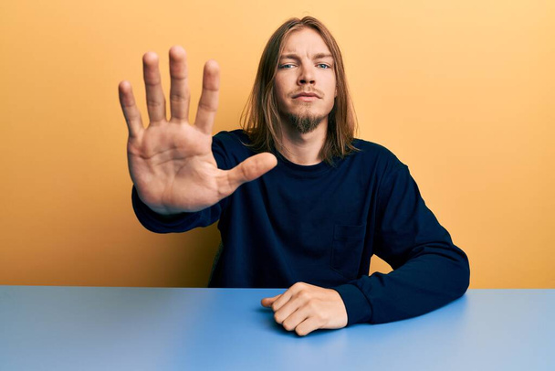 Handsome caucasian man with long hair wearing casual clothes sitting on the table doing stop sing with palm of the hand. warning expression with negative and serious gesture on the face.  - Fotografie, Obrázek