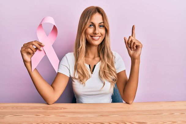 Beautiful blonde young woman holding pink cancer ribbon surprised with an idea or question pointing finger with happy face, number one  - Foto, immagini