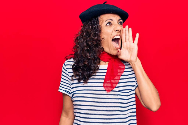 Middelbare leeftijd mooie vrouw draagt Franse baret schreeuwen en schreeuwen luid van kant met de hand op de mond. communicatieconcept.  - Foto, afbeelding