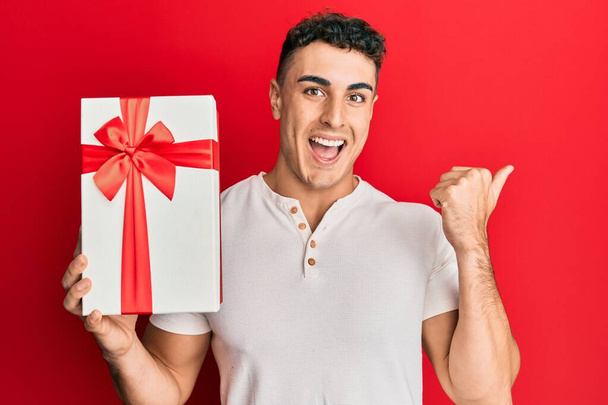 Іспанський молодий чоловік тримає подарунок, показуючи великий палець на стороні посміхаючись щасливий відкритим ротом  - Фото, зображення