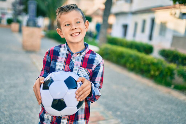 Αξιολάτρευτο καυκάσιο αγόρι χαμογελά χαρούμενος κρατώντας μπάλα ποδοσφαίρου στέκεται στο πάρκο. - Φωτογραφία, εικόνα