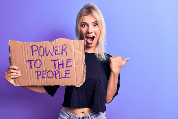 Junge blonde Frau, die für soziale Rechte eintritt, hält Banner mit Macht zum Volk und zeigt den Daumen zur Seite, lächelt glücklich mit geöffnetem Mund - Foto, Bild