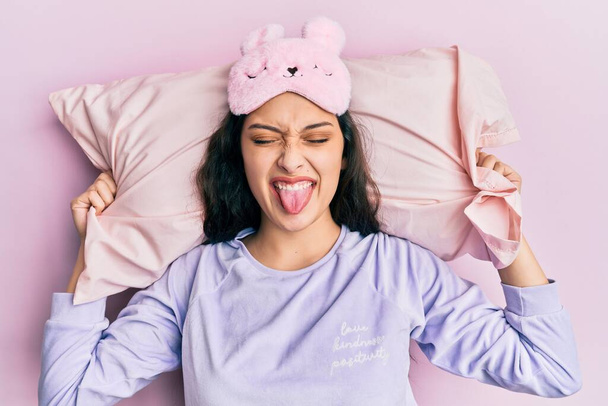 Hermosa mujer de Oriente Medio con máscara de sueño y pijama durmiendo en la almohada sacando la lengua feliz con expresión divertida.  - Foto, imagen