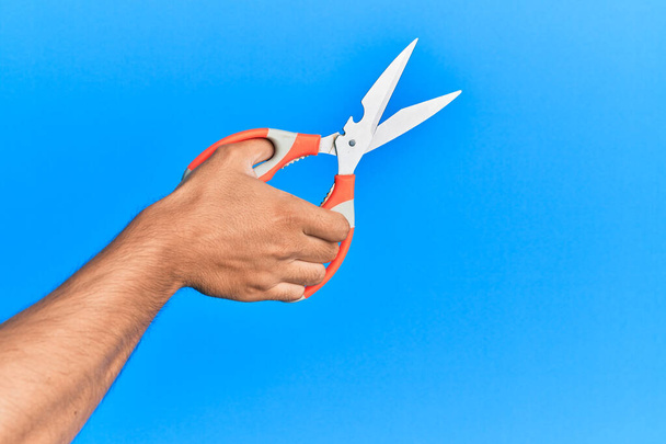 Hand of young hispanic man using scissors over isolated blue background. - Photo, Image