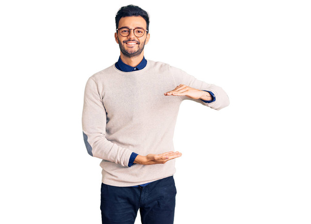 Junger gut aussehender hispanischer Mann in eleganter Kleidung und Brille gestikuliert mit Händen, die ein großes und großes Zeichen, ein Maßsymbol, zeigen. Lächelnd in die Kamera blickend. Messkonzept.  - Foto, Bild