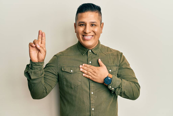 Jovem latino vestindo roupas casuais sorrindo jurando com a mão no peito e dedos para cima, fazendo uma promessa de lealdade juramento  - Foto, Imagem