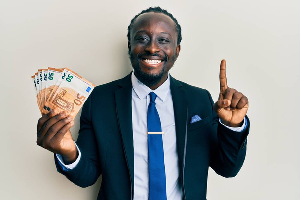 50ユーロ紙幣を保持するビジネススーツを身に着けているハンサムな若い黒人男性幸せな顔をしたアイデアや質問指差し指で笑顔,ナンバーワン  - 写真・画像