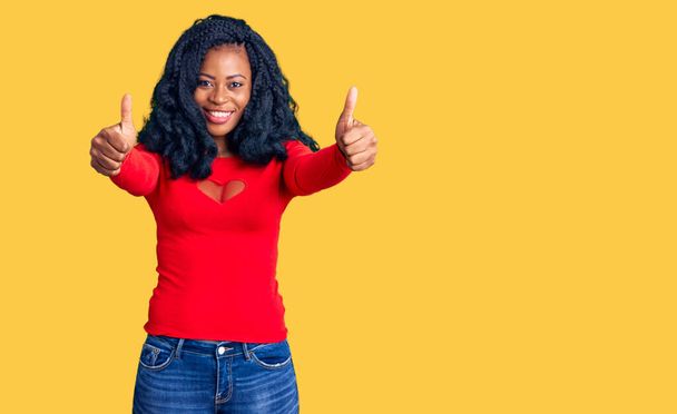 Krásná africká Američanka v neformálním oblečení, která schvaluje pozitivní gesto rukou, palce nahoru, usmívá se a je šťastná za úspěch. gesto vítěze.  - Fotografie, Obrázek