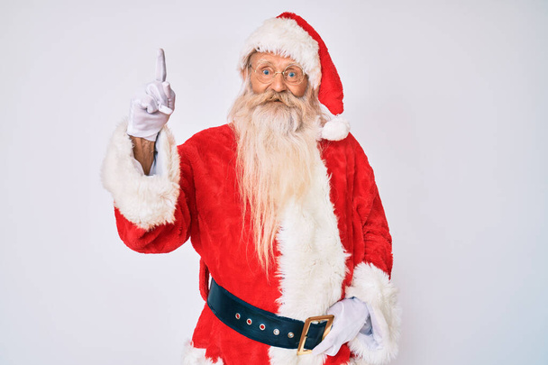 Old senior man with grey hair and long beard wearing santa claus costume with suspenders showing and pointing up with finger number one while smiling confident and happy.  - Φωτογραφία, εικόνα