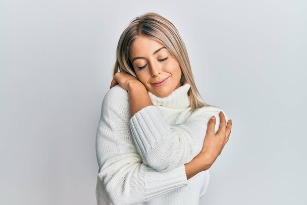 Beautiful blonde woman wearing casual turtleneck sweater hugging oneself happy and positive, smiling confident. self love and self care  - Foto, Imagem