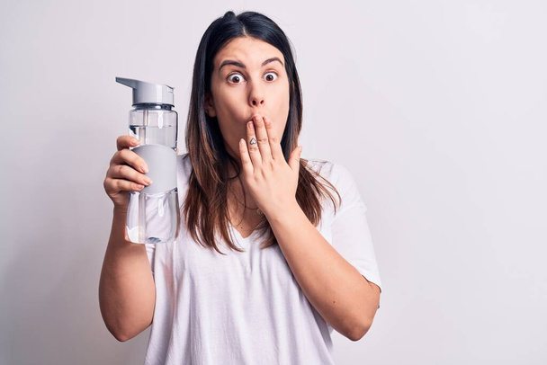 Mladá krásná brunetka žena pije láhev vody přes izolované bílé pozadí zakrývající ústa s rukou, šokovaný a strach z omylu. Překvapený výraz - Fotografie, Obrázek