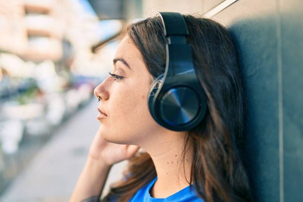 Mujer hispana joven con expresión seria escuchando música usando auriculares en la ciudad. - Foto, Imagen