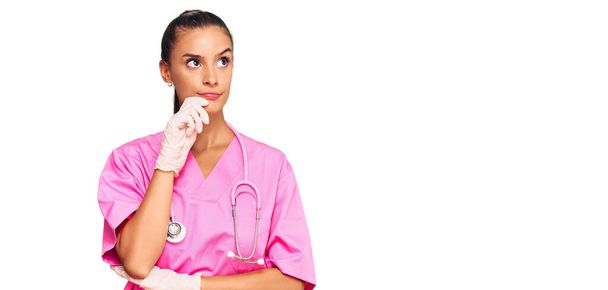 Young hispanic woman wearing doctor uniform and stethoscope serious face thinking about question with hand on chin, thoughtful about confusing idea  - Foto, Bild