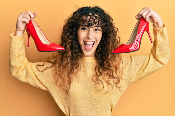 Chica hispana joven sosteniendo zapatos rojos de tacón alto celebrando loco y sorprendido por el éxito con los ojos abiertos gritando emocionado.  - Foto, Imagen