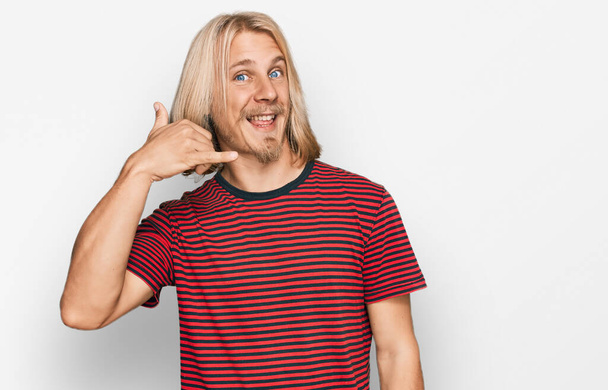 Homme caucasien aux cheveux longs blonds portant un t-shirt rayé décontracté souriant faisant geste téléphonique avec la main et les doigts comme parler au téléphone. communiquer les concepts.  - Photo, image