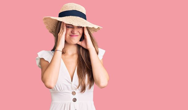 Junge schöne blonde Frau in Sommerkleid und Hut mit Kopfschmerzen verzweifelt und gestresst, weil Schmerzen und Migräne. Hände auf den Kopf.  - Foto, Bild