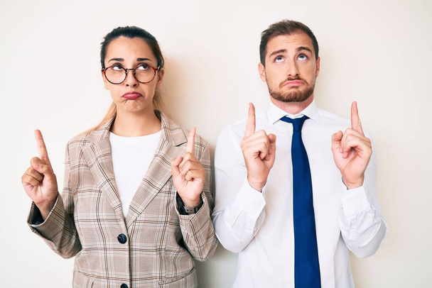 Beautiful couple wearing business clothes pointing up looking sad and upset, indicating direction with fingers, unhappy and depressed.  - Foto, Imagem