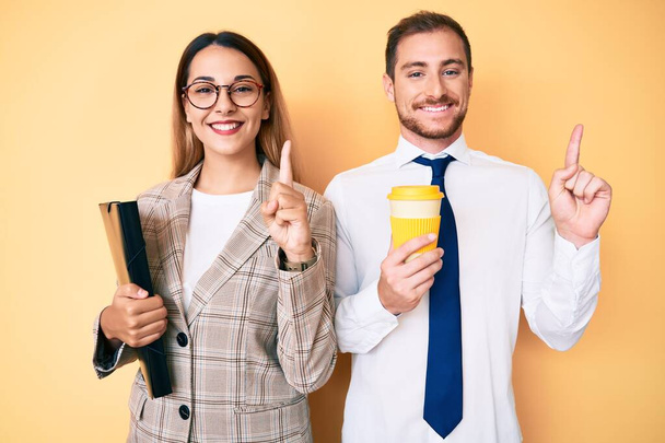 Beautiful couple wearing business clothes drinking take away coffee holding folder surprised with an idea or question pointing finger with happy face, number one  - Photo, Image