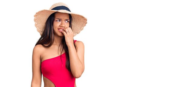 Junge schöne lateinamerikanische Mädchen in Badebekleidung und Sommermütze sehen gestresst und nervös aus, die Hände auf den Mund beißen Nägel. Angstproblem.  - Foto, Bild