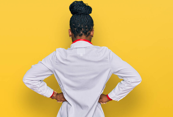 Νεαρή Αφροαμερικανή γυναίκα που φοράει ιατρική στολή και στηθοσκόπιο και στέκεται προς τα πίσω κοιτάζοντας μακριά με τα χέρια στο σώμα  - Φωτογραφία, εικόνα