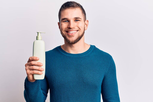 Young handsome man holding bottle of cream looking positive and happy standing and smiling with a confident smile showing teeth  - Photo, Image