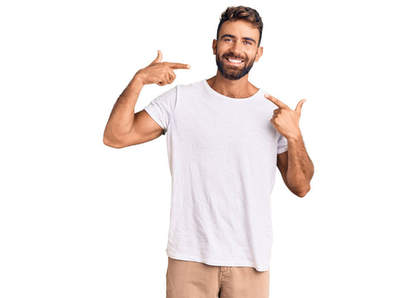 カジュアルな白いTシャツを着た若いヒスパニック系の男性は、陽気な笑みを浮かべて指の歯と口を指しています。歯の健康の概念.  - 写真・画像