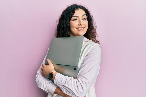 Junge brünette Frau mit lockigem Haar umarmt Laptop mit Liebe zwinkert in die Kamera mit sexy Ausdruck, fröhliches und glückliches Gesicht.  - Foto, Bild