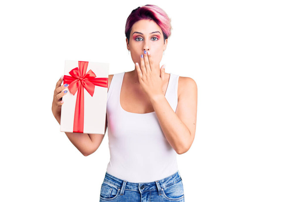 Νεαρή όμορφη γυναίκα με ροζ μαλλιά κρατώντας δώρο που καλύπτει το στόμα με το χέρι, σοκαρισμένος και φοβισμένος για λάθος. έκπληκτη έκφραση  - Φωτογραφία, εικόνα