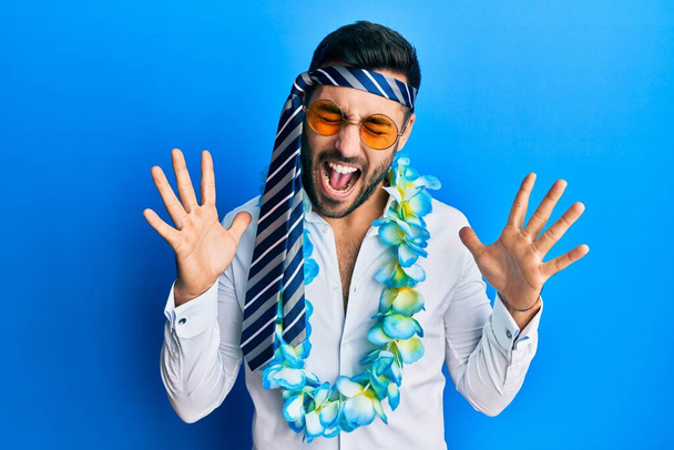 Junger hispanischer Geschäftsmann in lustiger Party-Manier mit Krawatte auf dem Kopf, der verrückt und erfolgsverrückt mit erhobenen Armen und geschlossenen Augen aufgeregt feiert. Siegerkonzept  - Foto, Bild