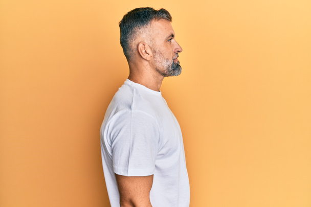Gut aussehender Mann mittleren Alters trägt lässiges weißes T-Shirt, das zur Seite schaut, entspannte Profilpose mit natürlichem Gesicht und selbstbewusstem Lächeln.  - Foto, Bild
