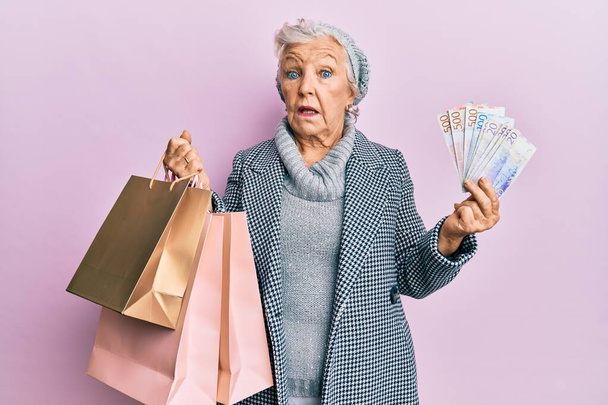 Idősebb ősz hajú nő, bevásárlószatyrokkal és svéd koronabankjegyekkel a kezében, tudatlan és zavaros arckifejezéssel. kétség.  - Fotó, kép